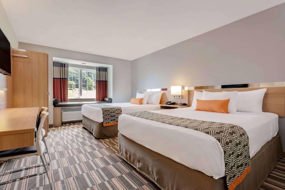 Habitación de hotel con dos camas, un escritorio, una silla, un ventanal con cortinas y vistas a zonas verdes.