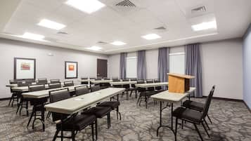 A conference room with multiple tables and chairs, a podium, framed pictures on the walls, and large windows with curtains.