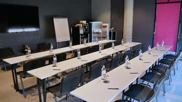 Ein Konferenzraum mit langen Tischen, Stühlen, einer Whiteboard-Tafel, einer Projektionsleinwand und einem Wasserspender.