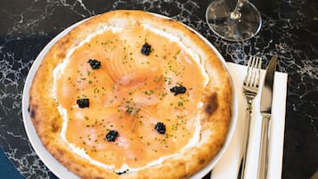 A round pizza with toppings on a white plate, accompanied by a fork and knife on a napkin, and a glass of white wine on a marble table.
