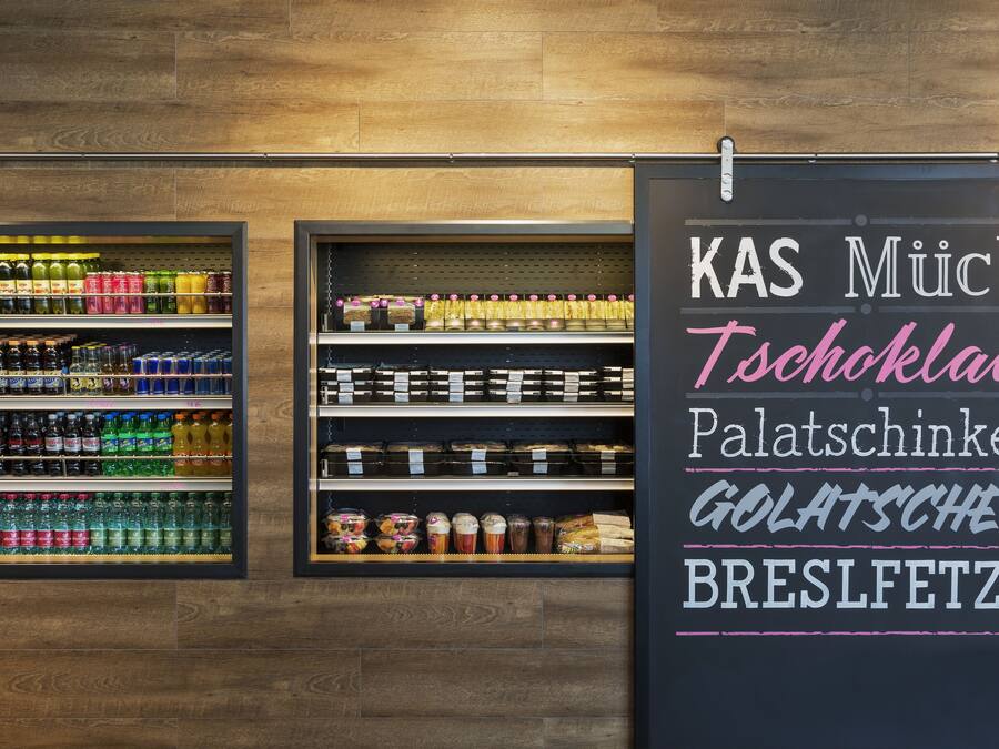 Un présentoir de boissons avec une variété de breuvages et un tableau noir affichant le menu, avec des articles tels que « KAS Much Tschoklad Palatschinken ».