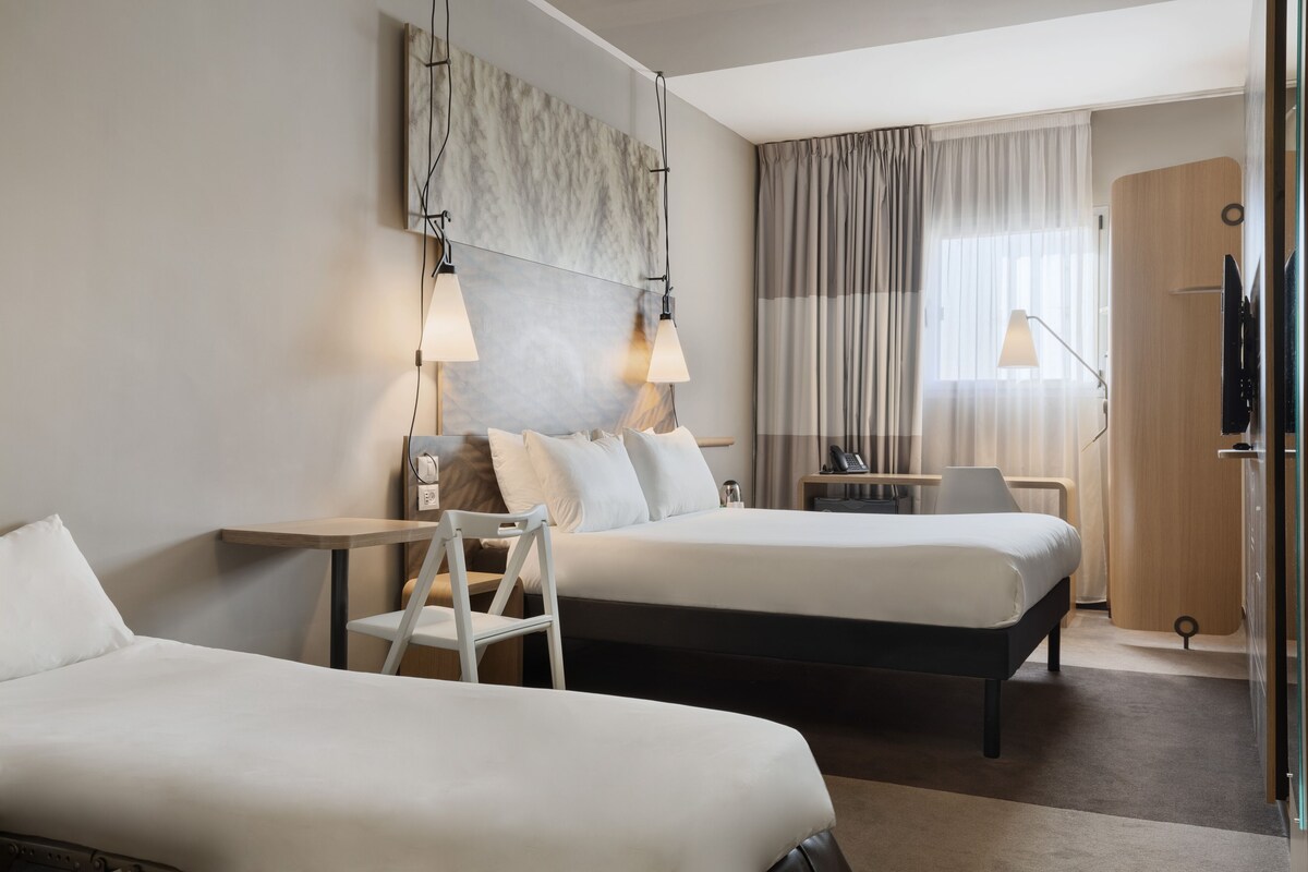 A hotel room with two beds, a desk, a chair, a TV, and a large window with curtains.