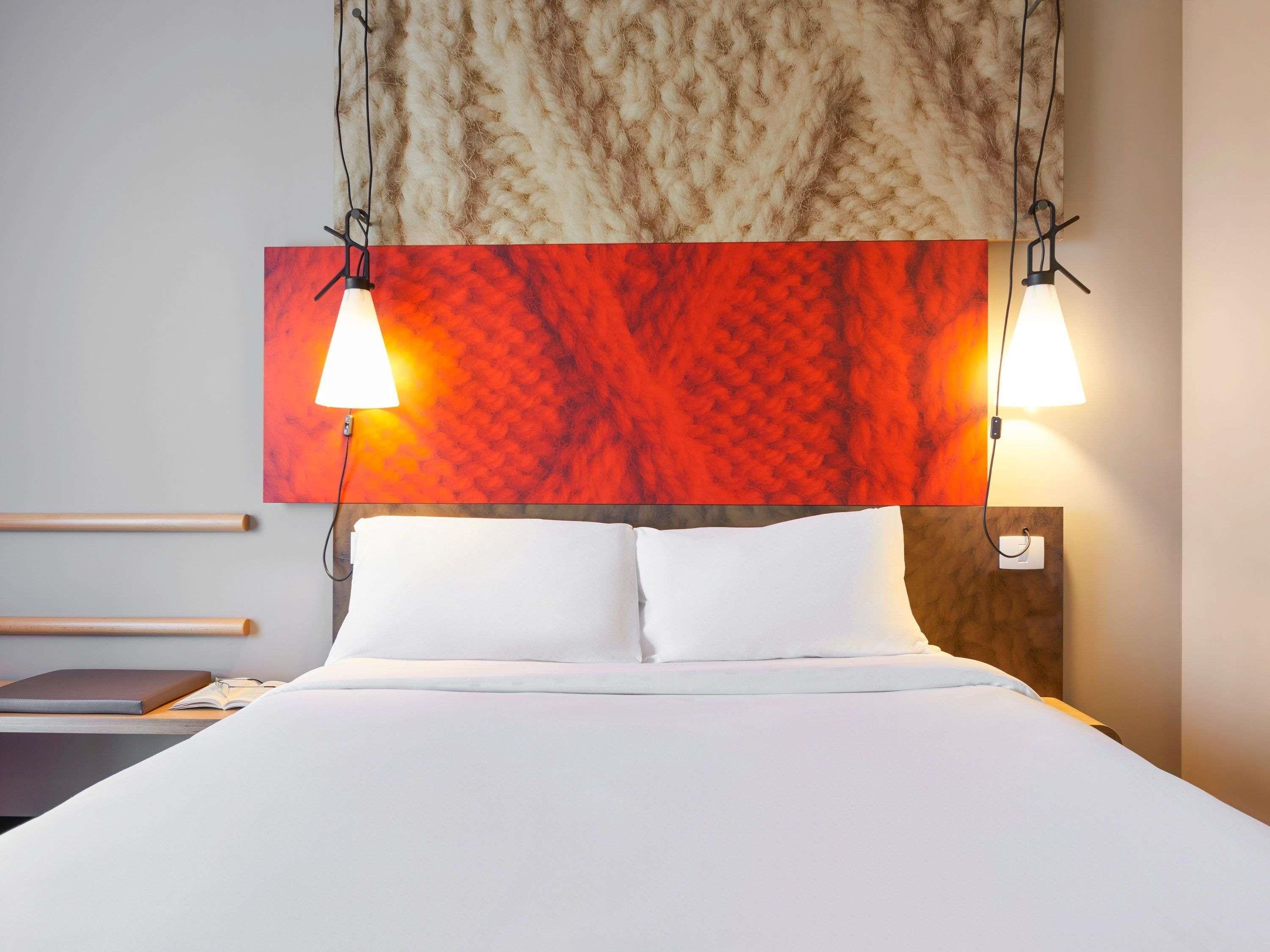 A hotel room with a bed, two bedside lamps, a headboard with a textured design, and a wall-mounted artwork.