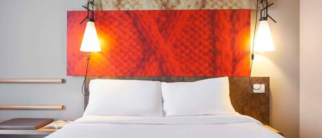 A hotel room with a bed, two bedside lamps, a headboard with a textured design, and a wall-mounted artwork.