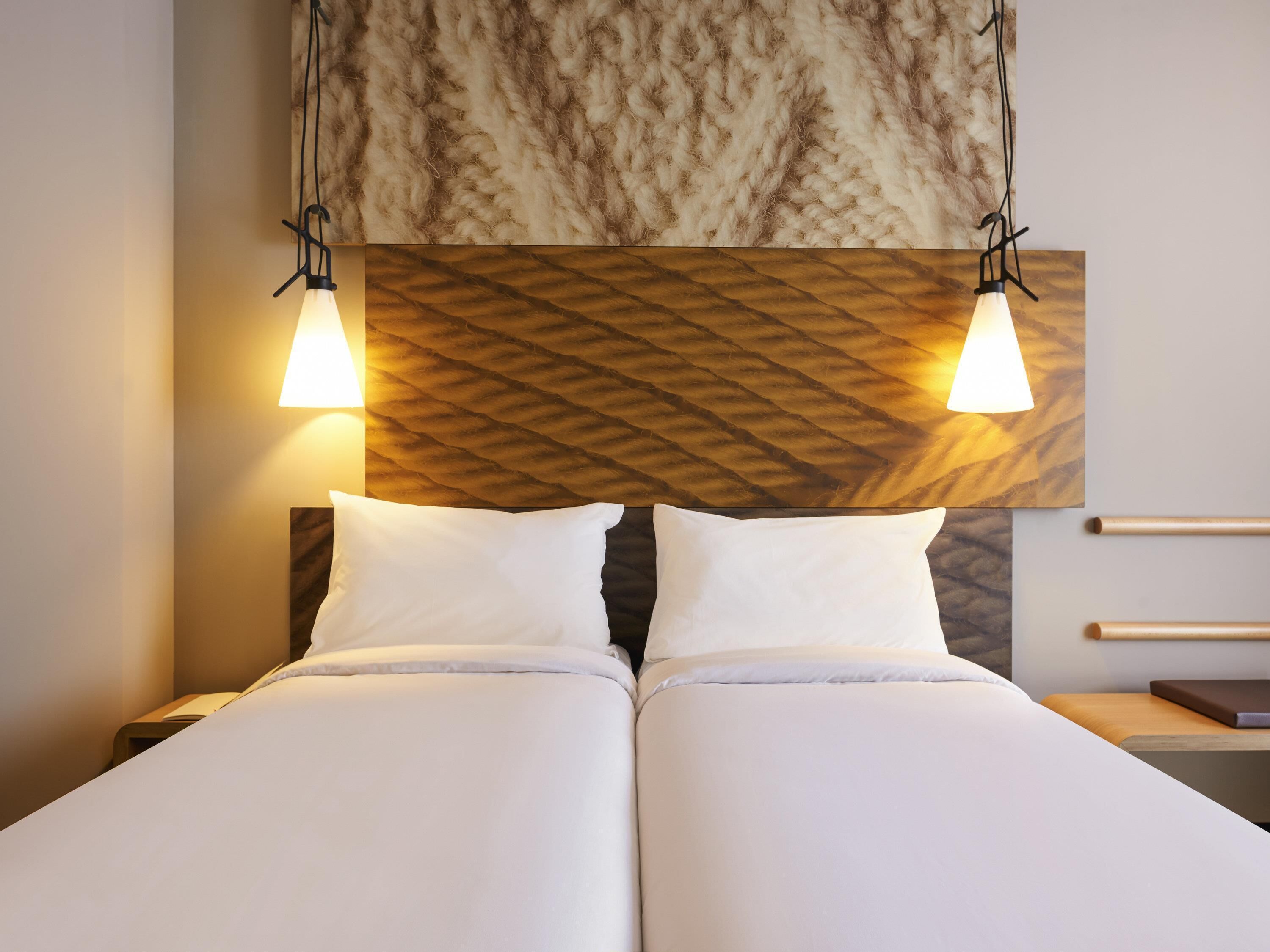 Un lit double avec du linge de lit blanc, deux tables de chevet, une tête de lit en bois ornée d’un motif géométrique et deux lampes suspendues.