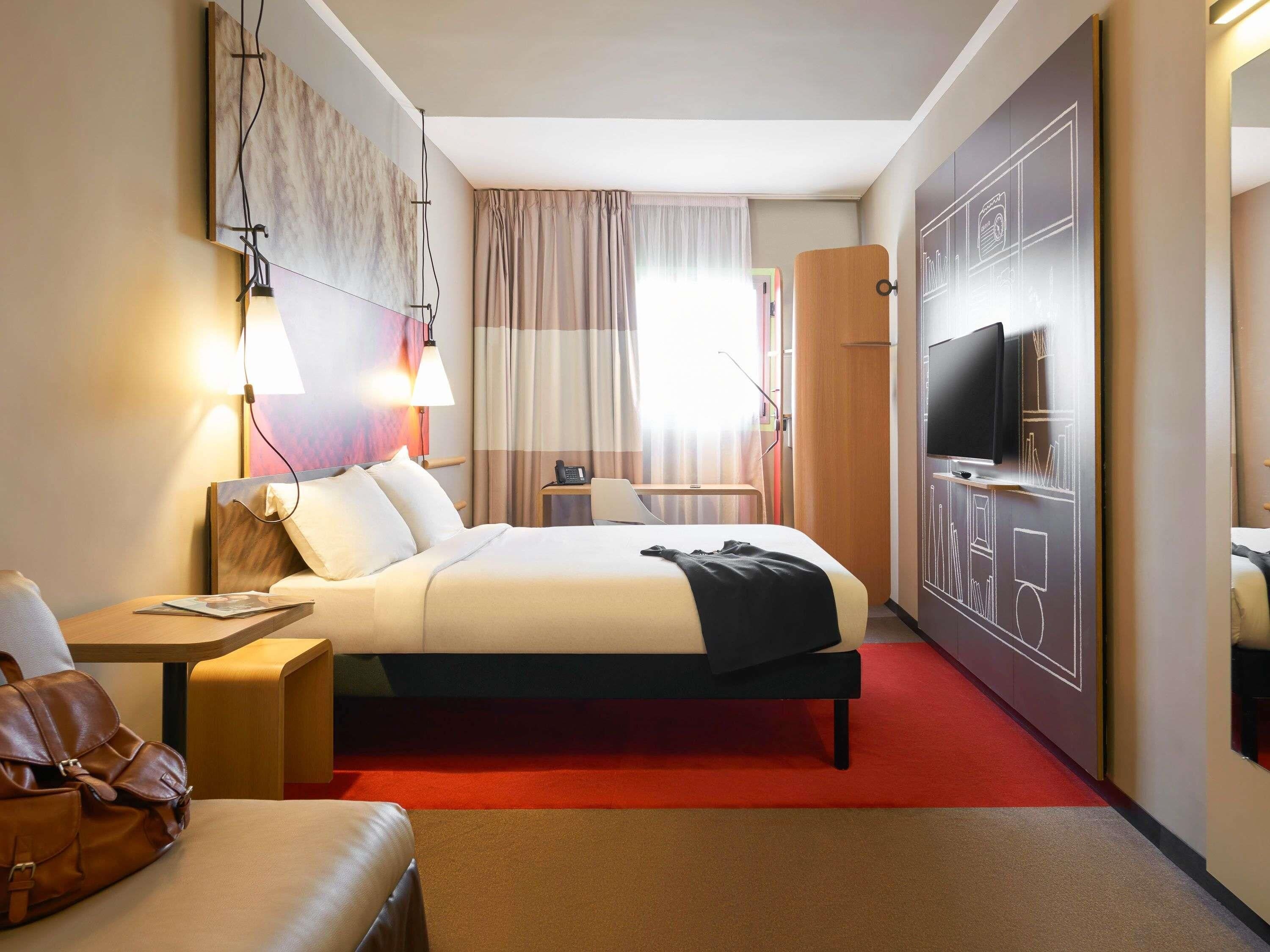 Quarto de hotel com cama, mesa de cabeceira, luminária, televisão e tapete vermelho.