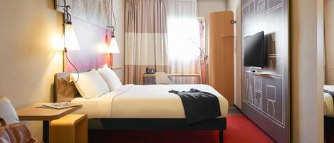 Quarto de hotel com cama, mesa de cabeceira, luminária, televisão e tapete vermelho.