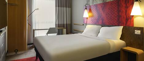 Una habitación de hotel con una cama grande, un escritorio, una silla y un ventanal con cortinas.