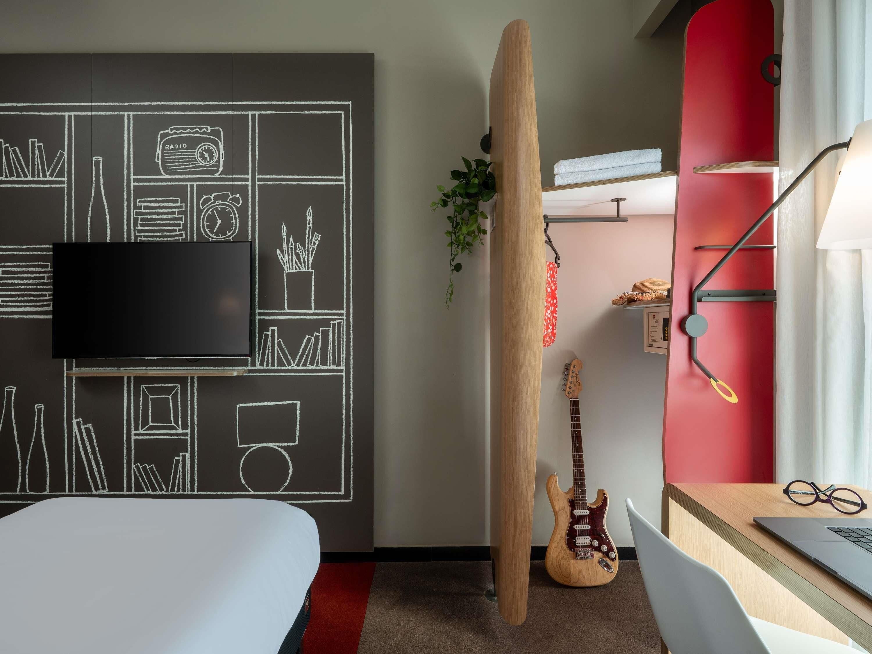 A room with a bed, a guitar, a surfboard, a TV, and a desk with a laptop and glasses.