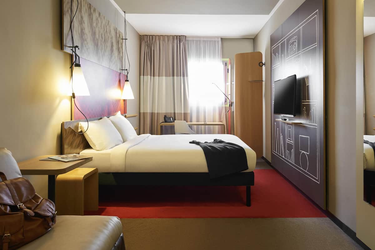 Una habitación de hotel con cama, mesita de noche, lámpara, televisor y alfombra roja.