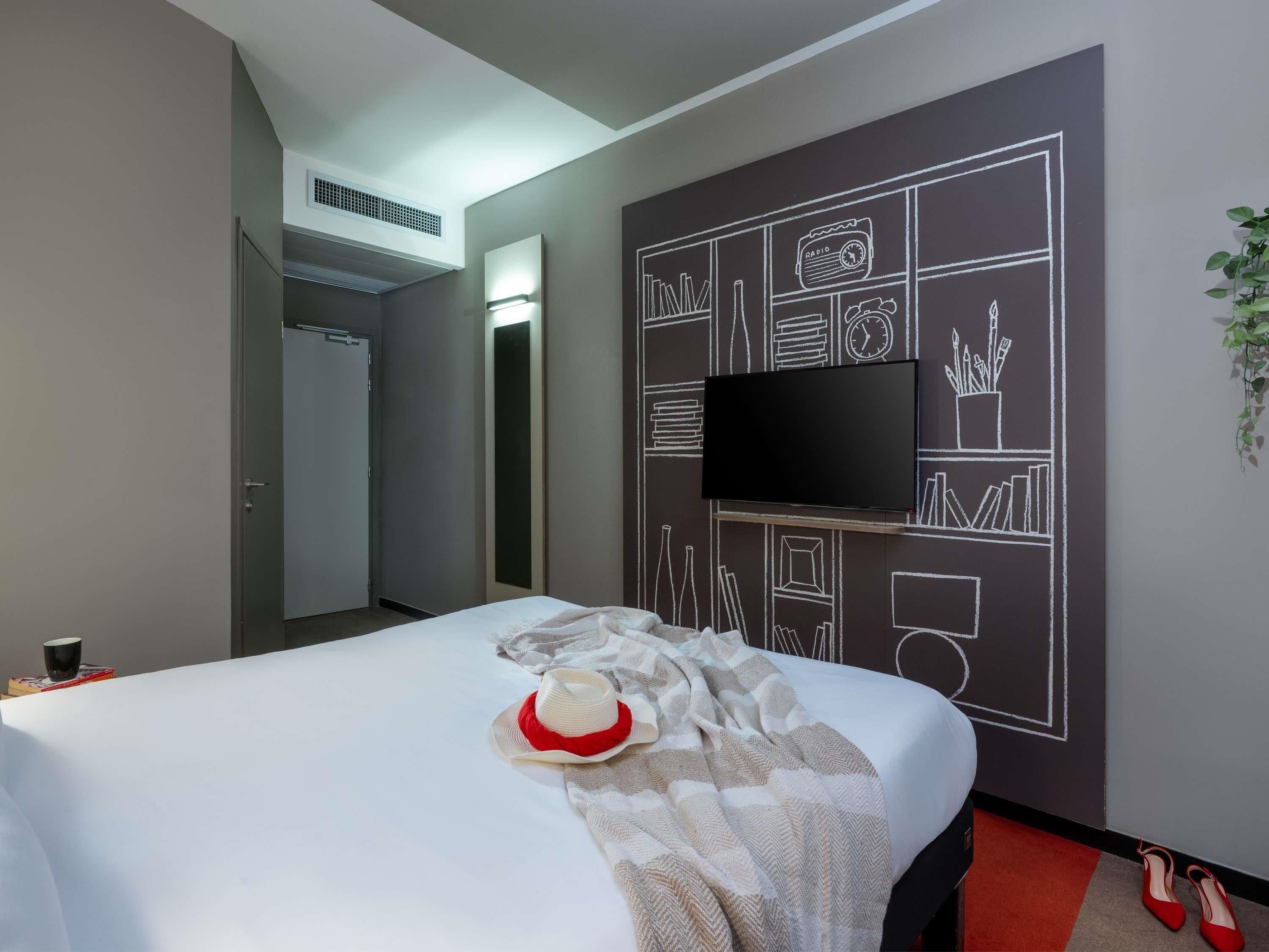 A hotel room with a bed, a flat-screen TV, a wardrobe, and a red carpet.