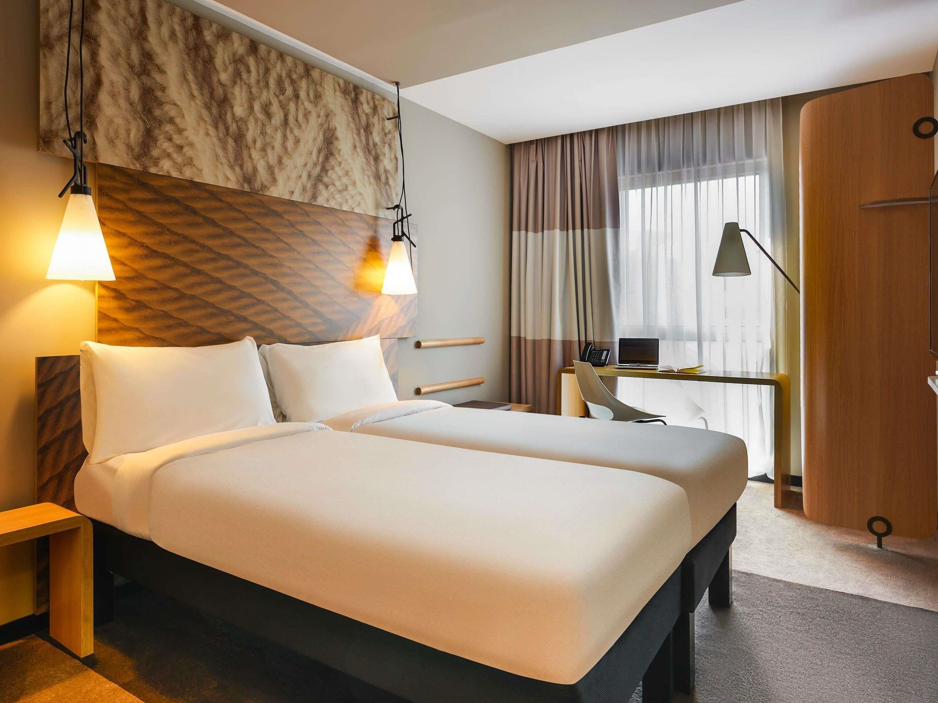 A hotel room with a large bed, bedside tables, a desk with a laptop, and a window with curtains.