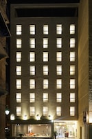 Un hotel de varios pisos con ventanas uniformemente iluminadas, una entrada a nivel de calle con mostrador de recepción y un área de estar exterior visible.