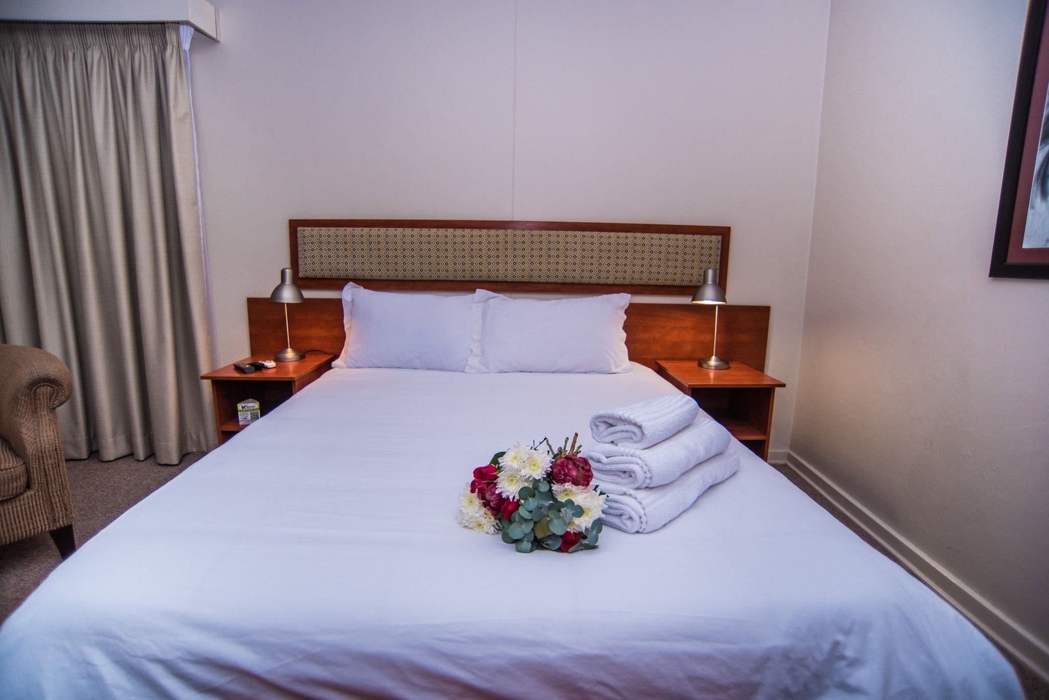Ein Hotelzimmer mit einem großen Bett, einem hölzernen Kopfteil, zwei Nachttischen mit Lampen, einem Sessel und einem Blumenstrauß auf dem Bett.
