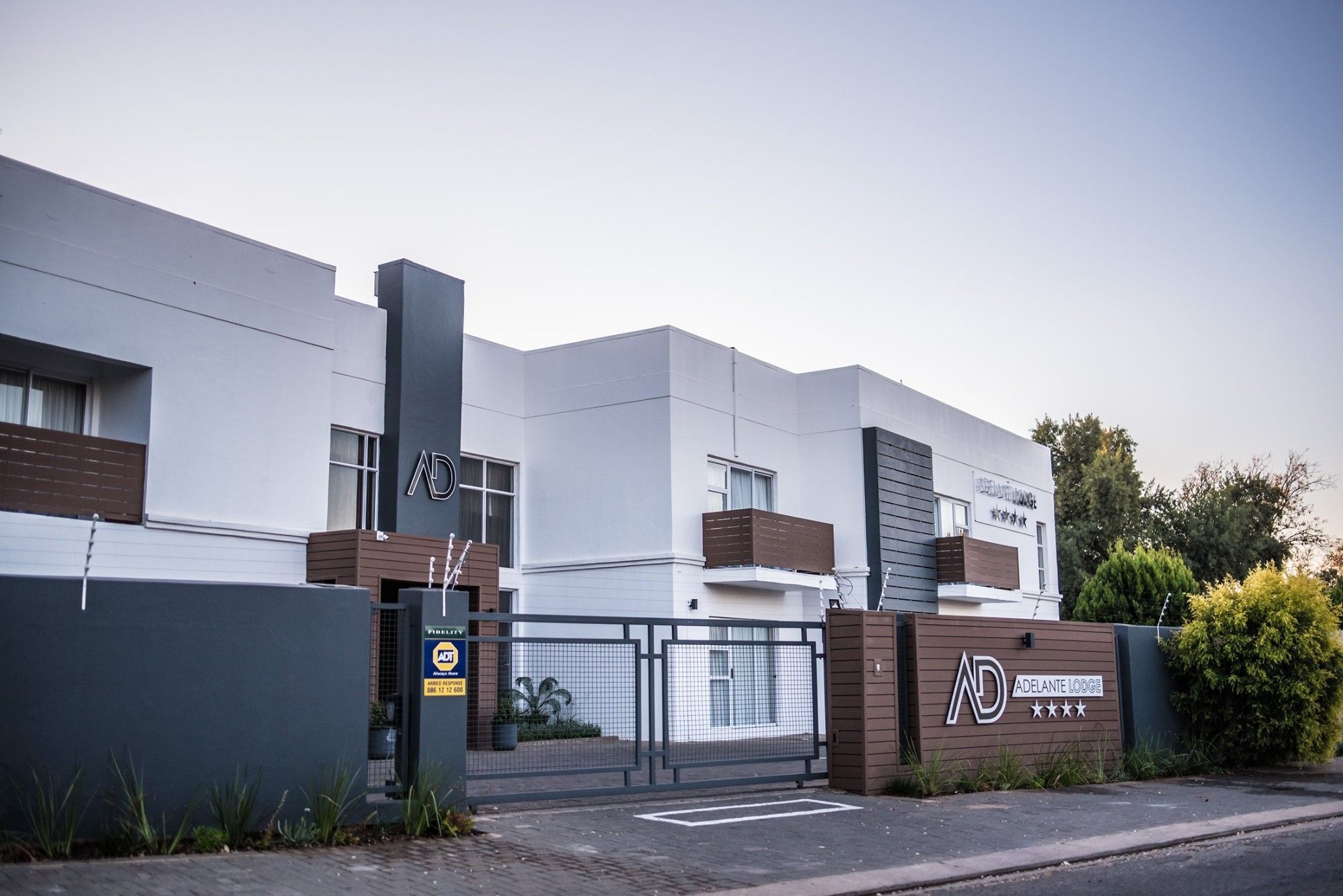 A modern building with a gated entrance, a sign for 'Adelante Lodge', and a clear sky.