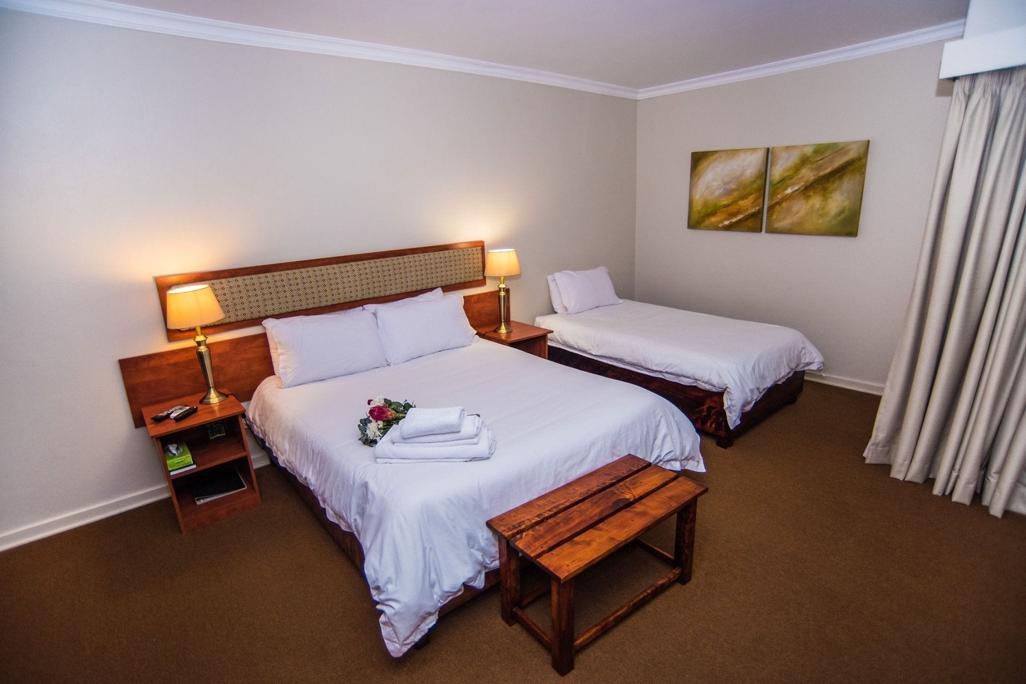 Una camera d'albergo con due letti, una panca di legno, un comodino con una lampada e un piccolo ripiano.
