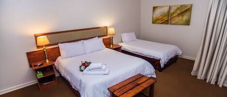 A hotel room with two beds, a wooden bench, a nightstand with a lamp, and a small shelf.