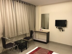 Una habitación con televisión, escritorio, dos sillas, una mesita de vidrio y una ventana con cortinas.