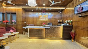A reception area with a wooden reception desk, a clock, a television, and a floral arrangement. - Hotel Jashn Inn near Apollo Hospital, Delhi (New Delhi)
