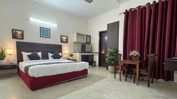 A bedroom with a bed, bedside tables, a desk, chairs, a dining table, a potted plant, and a red curtain. - Hotel Jashn Inn near Apollo Hospital, Delhi (New Delhi)