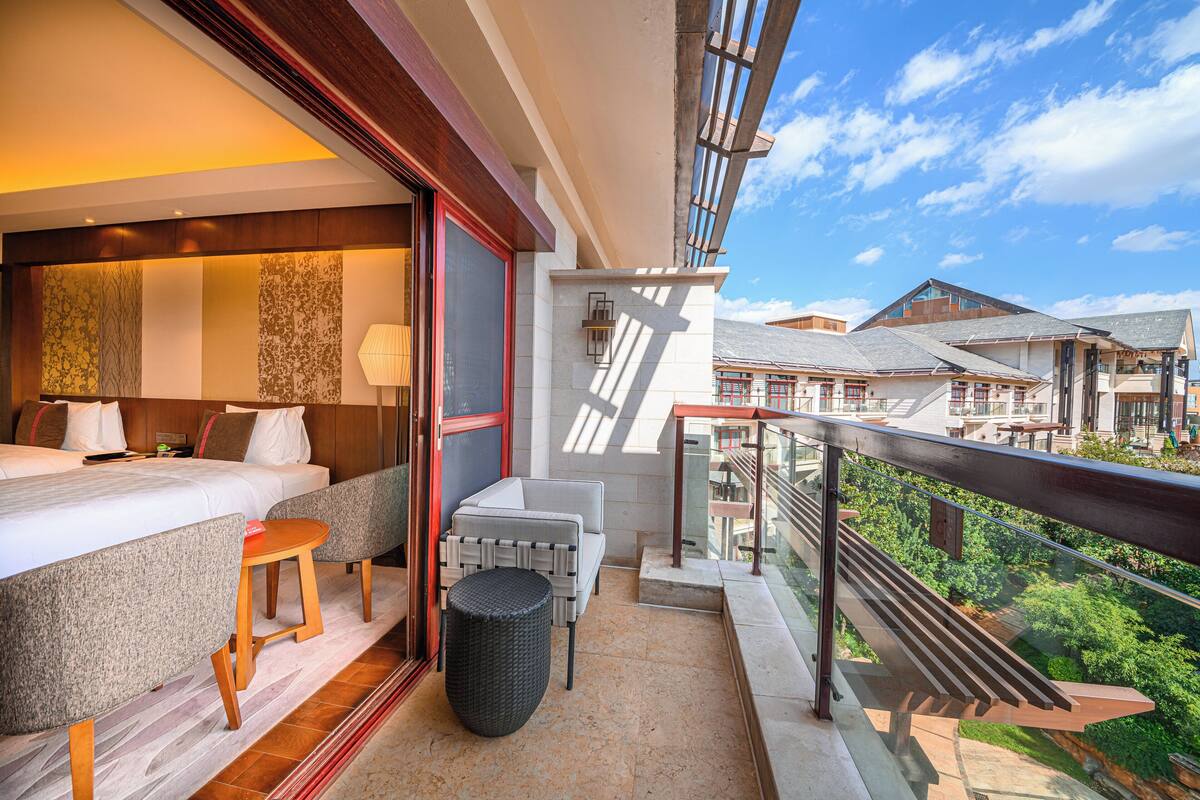 A balcony with a view of a mountainous landscape, a bed with a nightstand, a chair, and a small table.