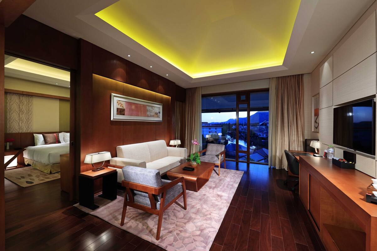 A modern hotel room with a large bed, a sofa, a coffee table, and a TV. The room has wooden flooring and a large window with a view of the mountains.
