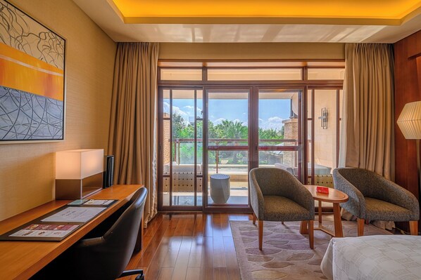 A hotel room with a wooden desk, two armchairs, a bed, and a large sliding glass door leading to an outdoor area with a view of greenery and buildings. - HUALUXE Kunming by IHG (Kunming)