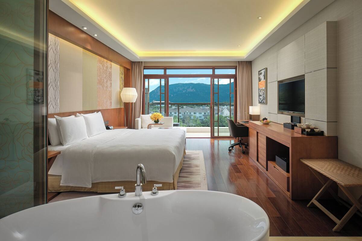 A modern hotel room with a large bed, a desk, a TV, and a view of mountains.