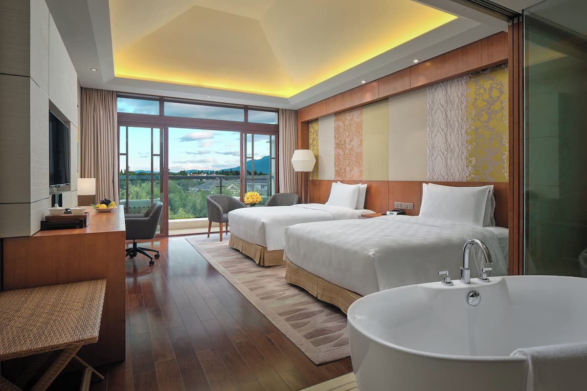 A hotel room with two beds, a large window, a TV, a desk, and a bathtub.