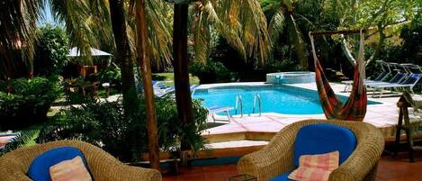 A poolside area with wicker chairs and a hammock, surrounded by lush greenery and a clear view of the pool.