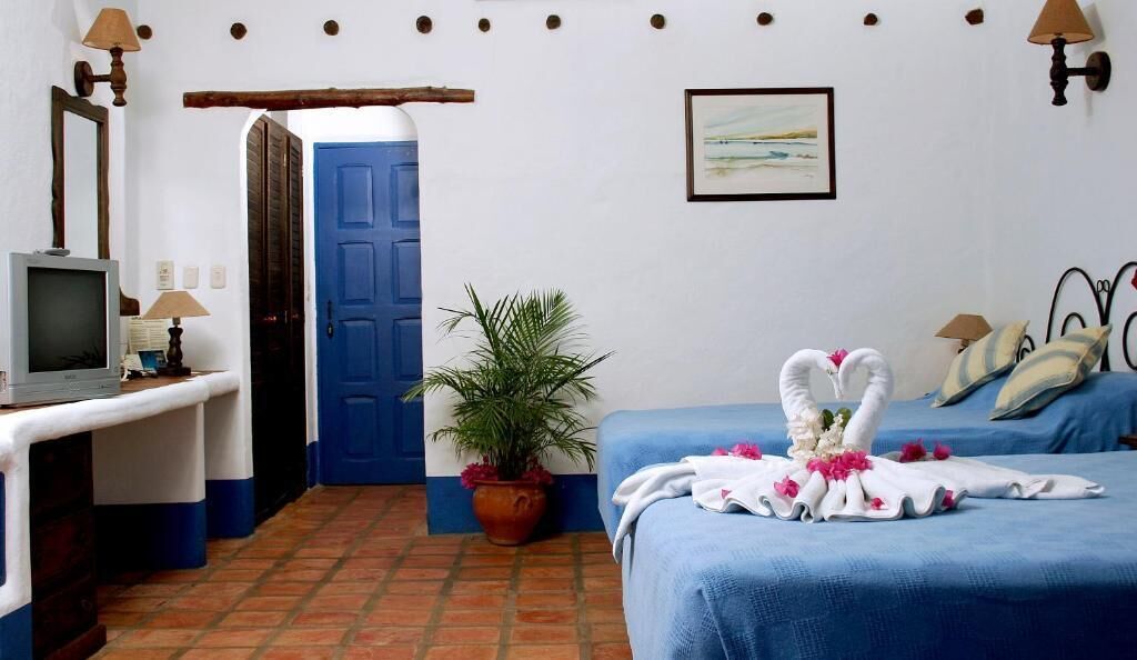 A room with a blue door, a desk with a lamp, a potted plant, and a bed with a towel and flowers.