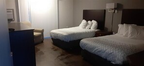 Two single beds with white linens and multiple pillows, a nightstand with a lamp, and a desk with a lamp.