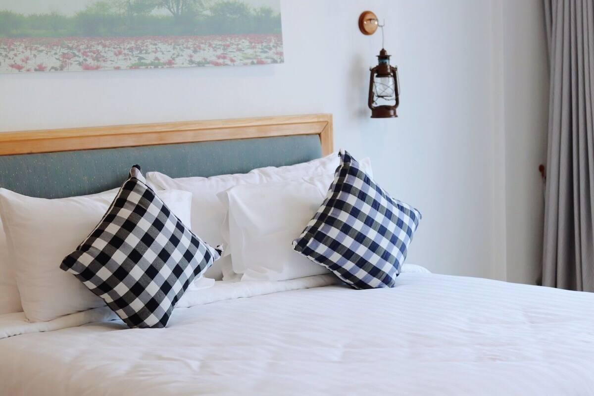 A bed with white bedding and two checkered pillows, a wooden headboard, a wall-mounted lantern, and a painting of a field.