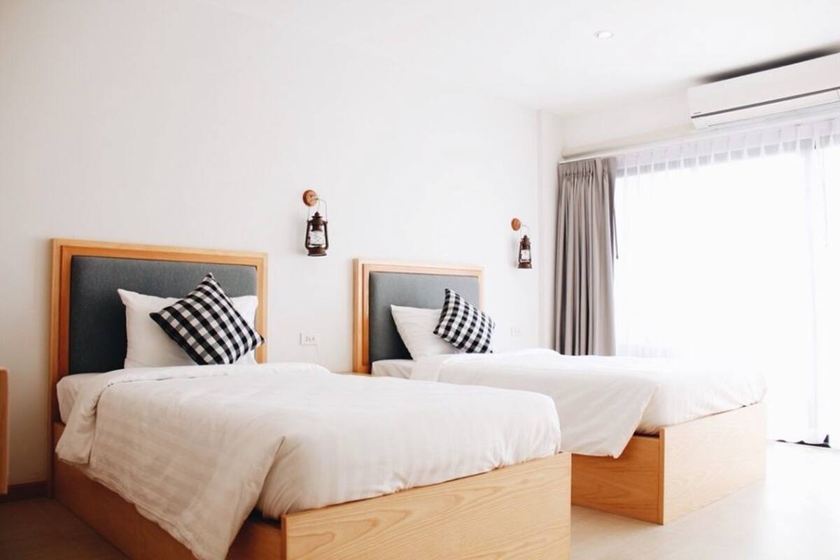 A hotel room with two single beds, each with a wooden headboard and white bedding. There are two wall-mounted lanterns and an air conditioning unit on the wall.