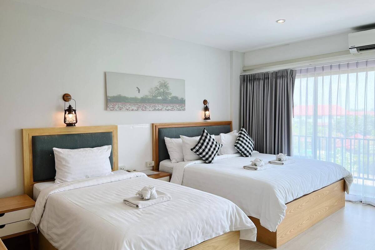 A hotel room with two beds, each with a wooden headboard and white bedding. There are two bedside tables with lamps, a painting on the wall, and a window with curtains.