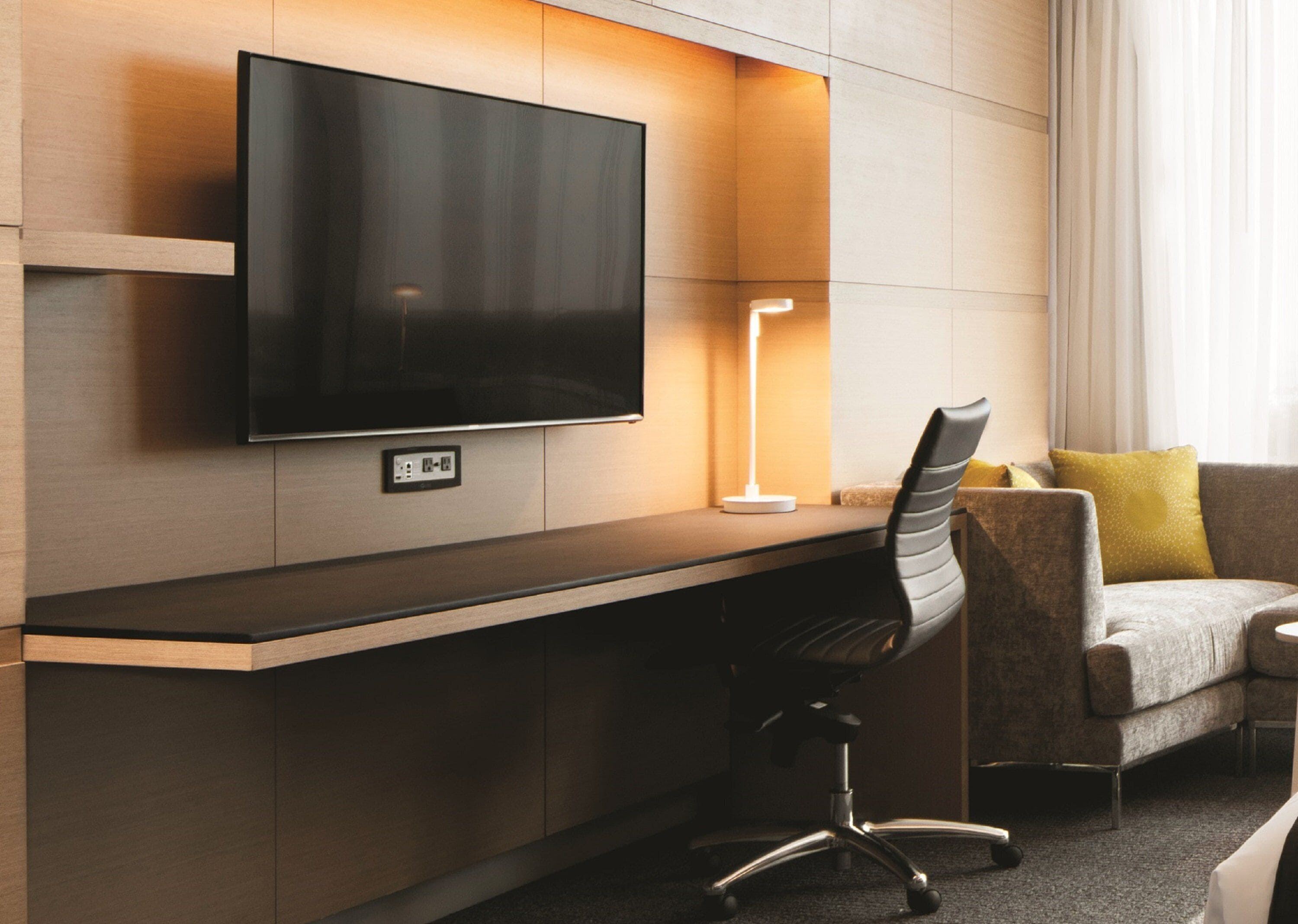 A modern hotel room with a flat-screen TV mounted on a wooden wall, a desk with a lamp, a cushioned armchair, and a sofa with a yellow pillow.