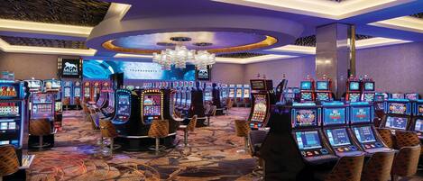 Un salón de casino con múltiples máquinas tragamonedas, un escenario con una pantalla grande y un techo decorativo.
