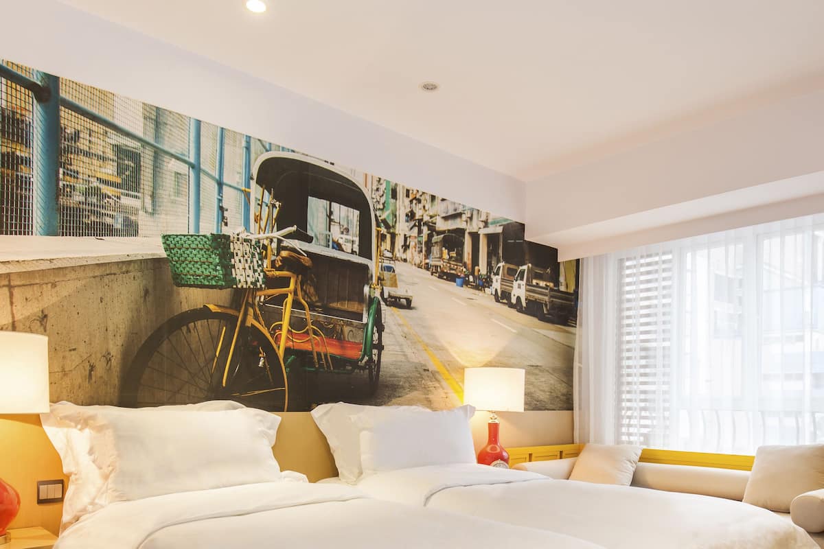 A hotel room with two beds, a large mural of a street scene, a window with blinds, and a nightstand with a lamp.