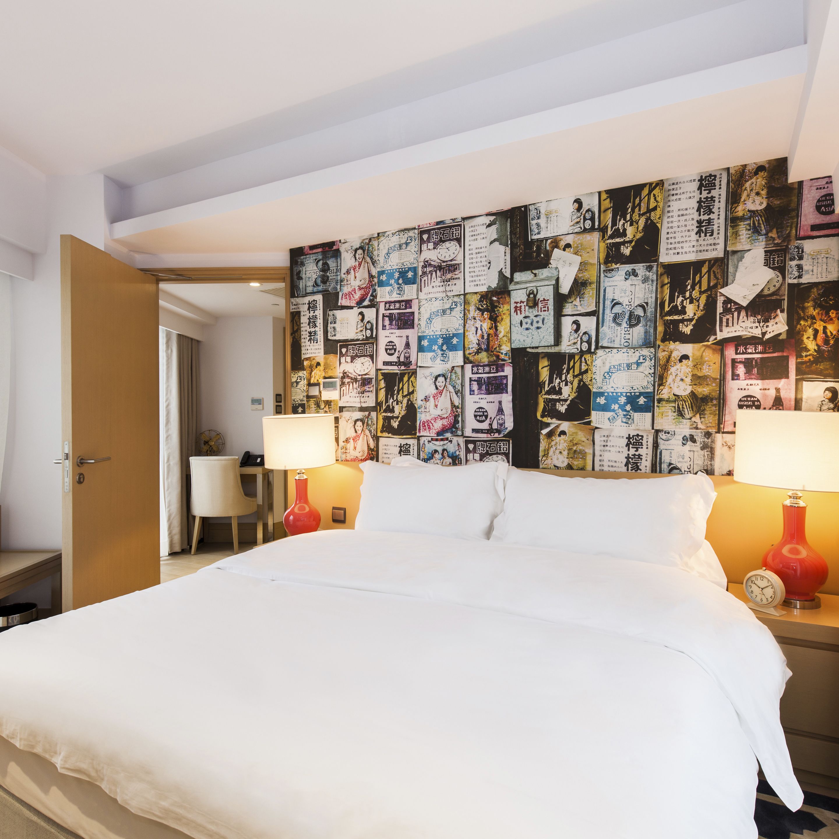 Una camera d'albergo con un letto grande, due comodini con lampade, un muro decorato con diversi poster e immagini, e una porta che conduce a un'altra stanza.