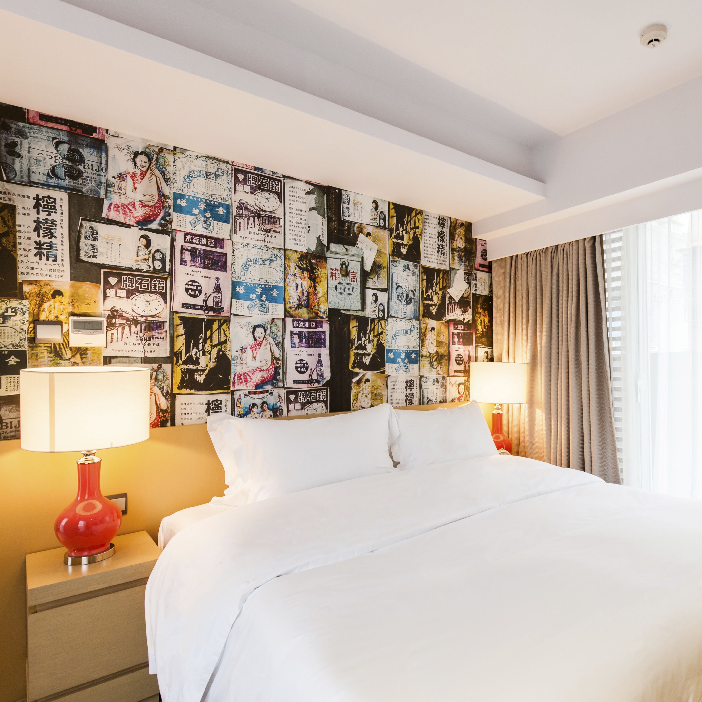 Une chambre d’hôtel avec un lit, deux tables de chevet, une lampe rouge et un mur recouvert de divers affiches.