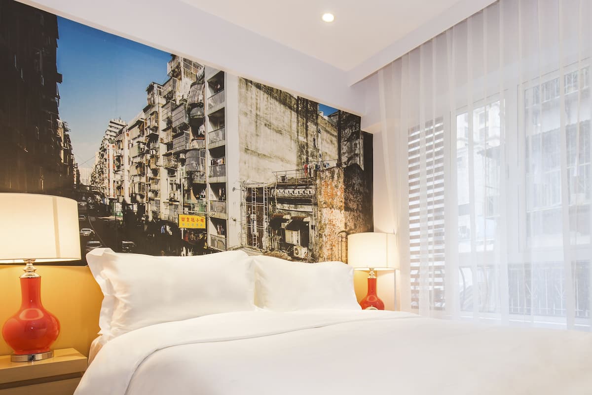 A hotel room with a large bed, two bedside tables with lamps, a cityscape mural on the wall, and a window with blinds.