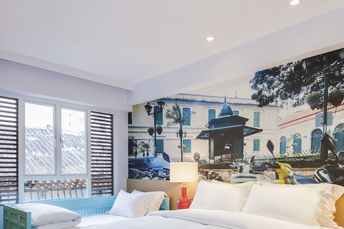A hotel room with a large bed, a window with blinds, a wall mural, and a small table with a lamp.