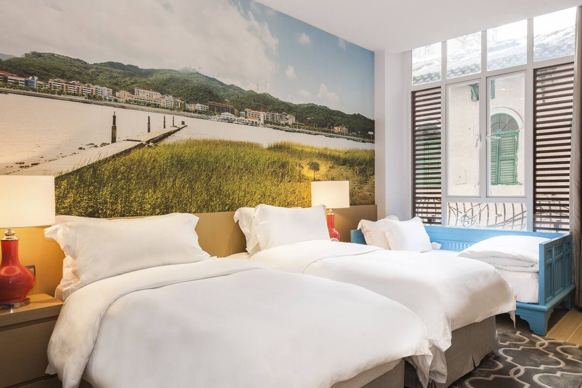 A hotel room with two beds, a large window with shutters, a mural of a landscape, and a patterned rug.