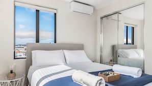 A hotel room with a large bed, a bedside table with a lamp, a window with a city view, and an air conditioning unit. - 110 Hampden Apartments (Battery Point)