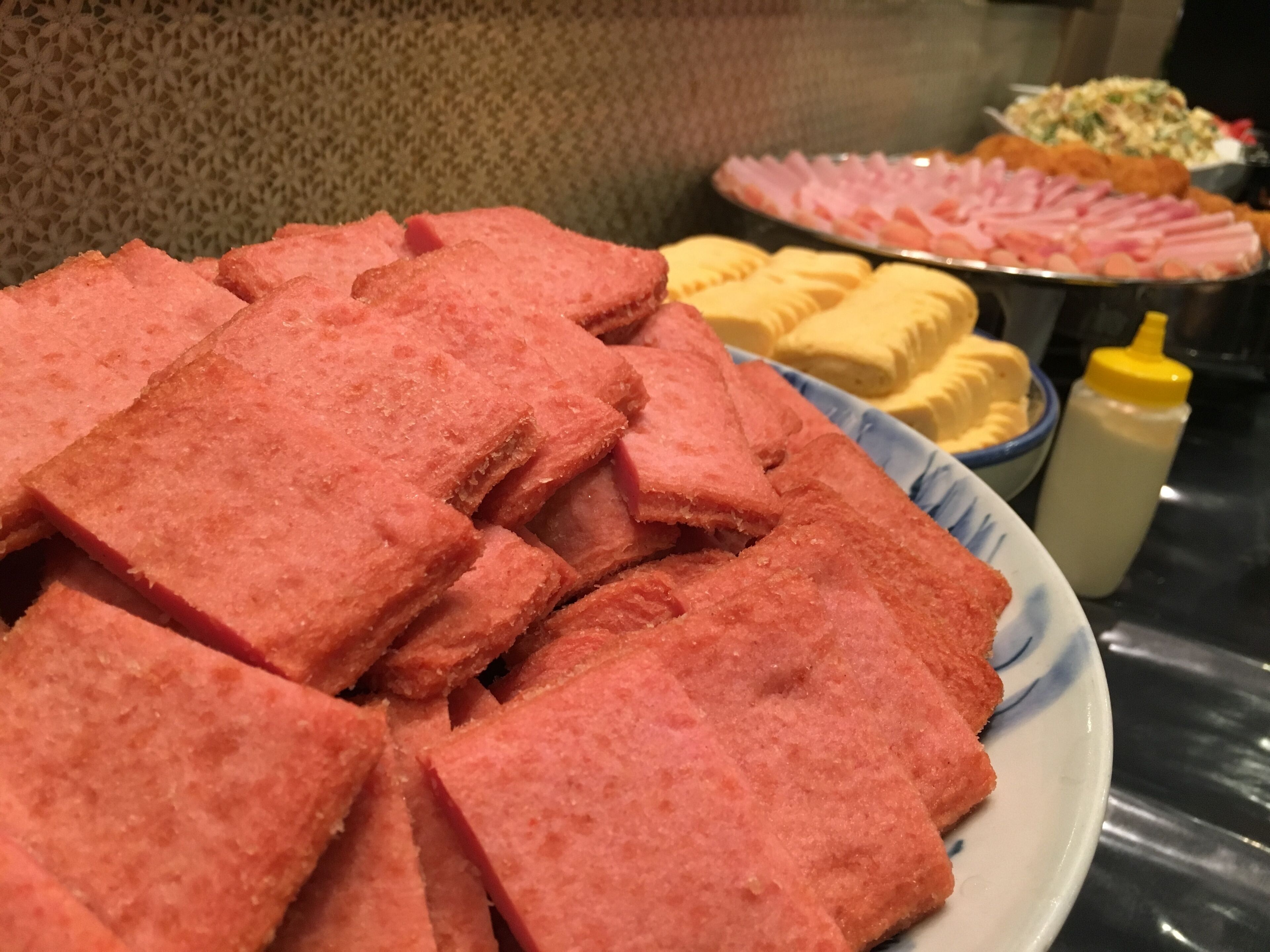 Une assiette contenant divers aliments roses, peut-être du pain, avec une bouteille de sauce jaune et un bol d’aliments jaunes en arrière-plan.