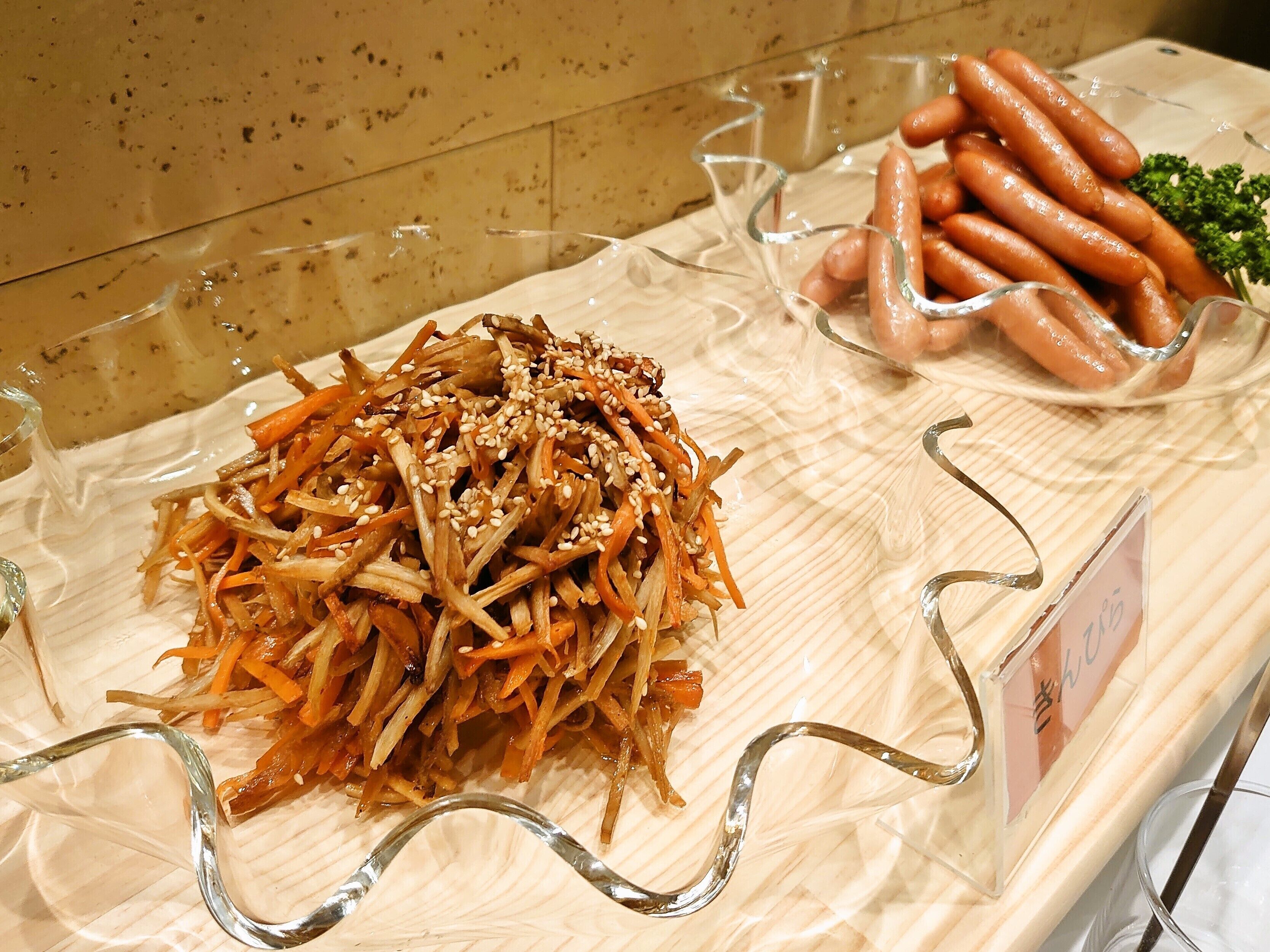 Eine Auswahl an Speisen, darunter eine Portion gehobelte Gemüse mit Sesam, eine Schüssel Würstchen und eine kleine Schüssel Salat.