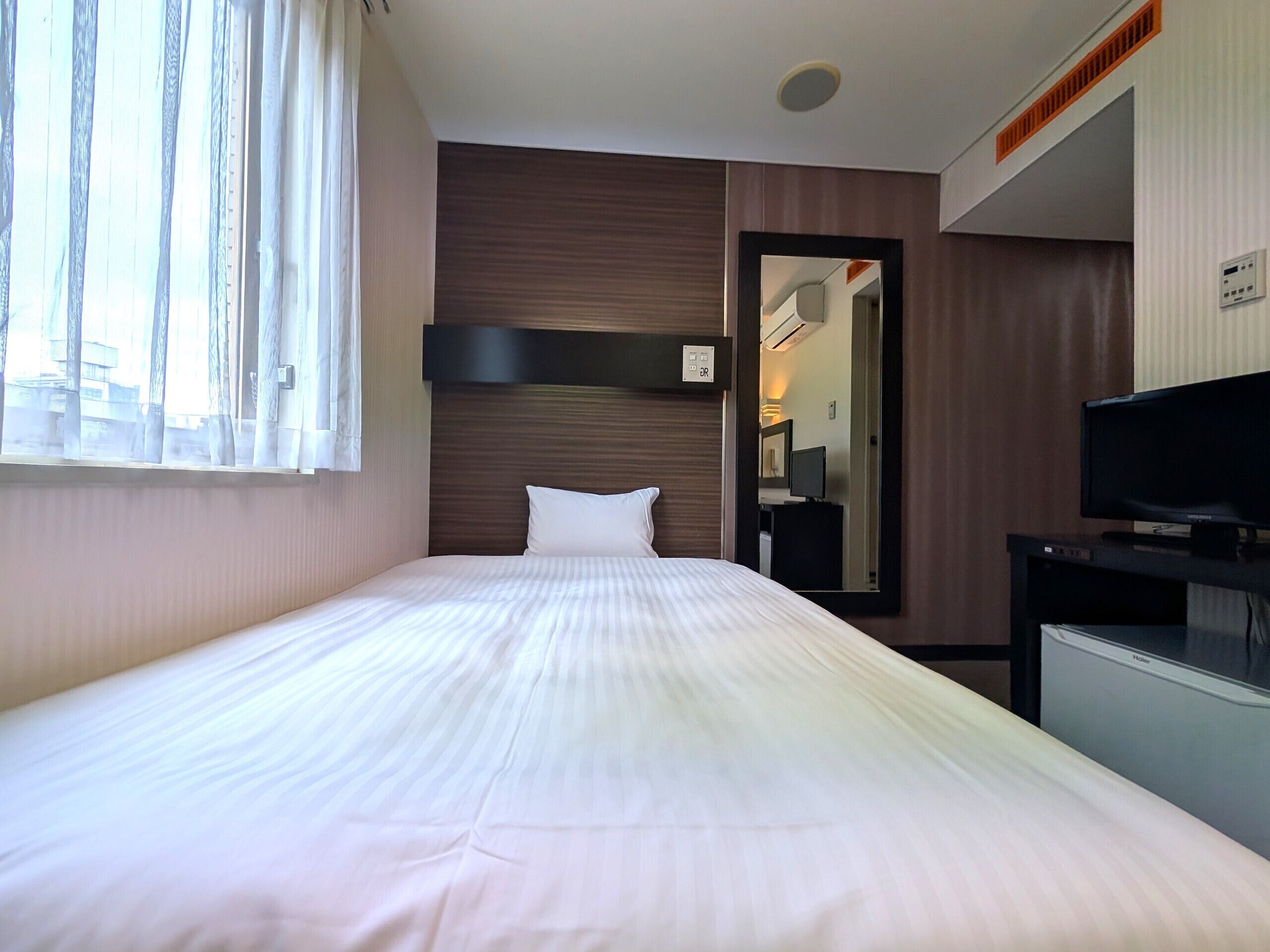 Habitación de hotel con cama, televisor, espejo y ventana con cortinas.