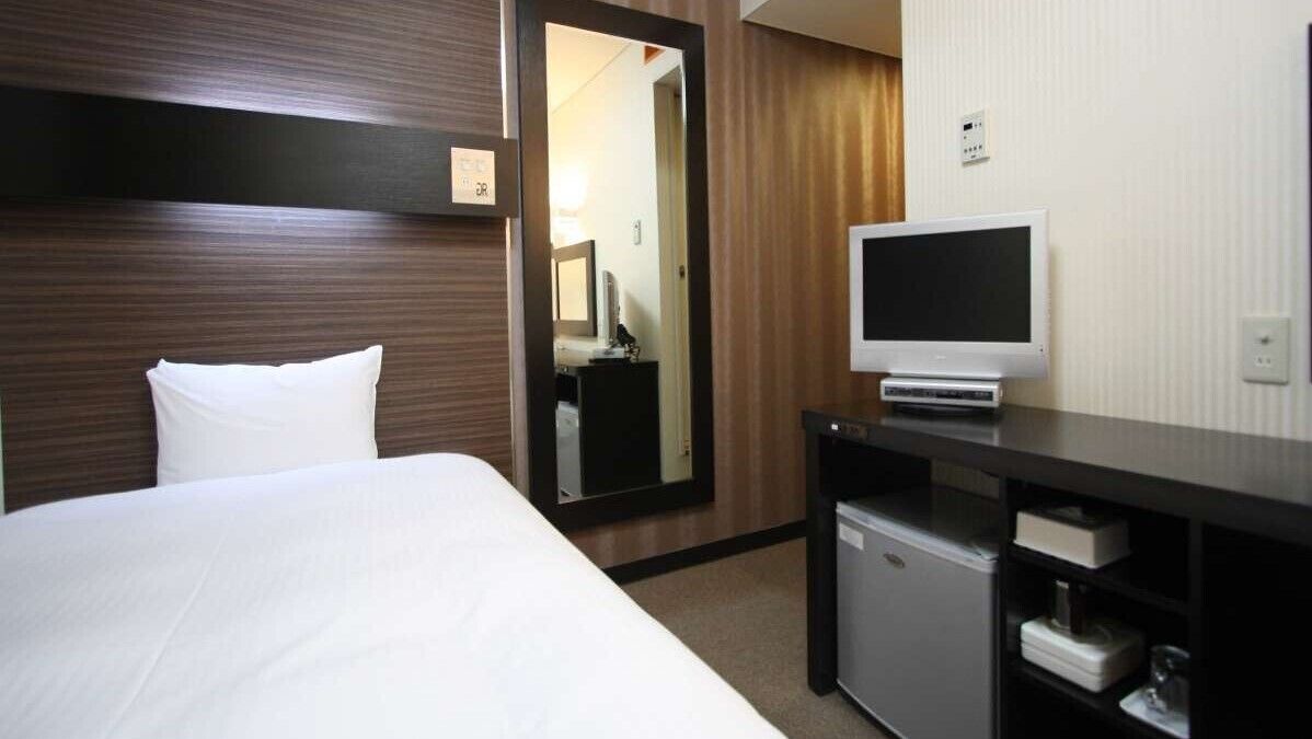 A hotel room with a bed, a small desk with a TV, a mirror, and a wardrobe.