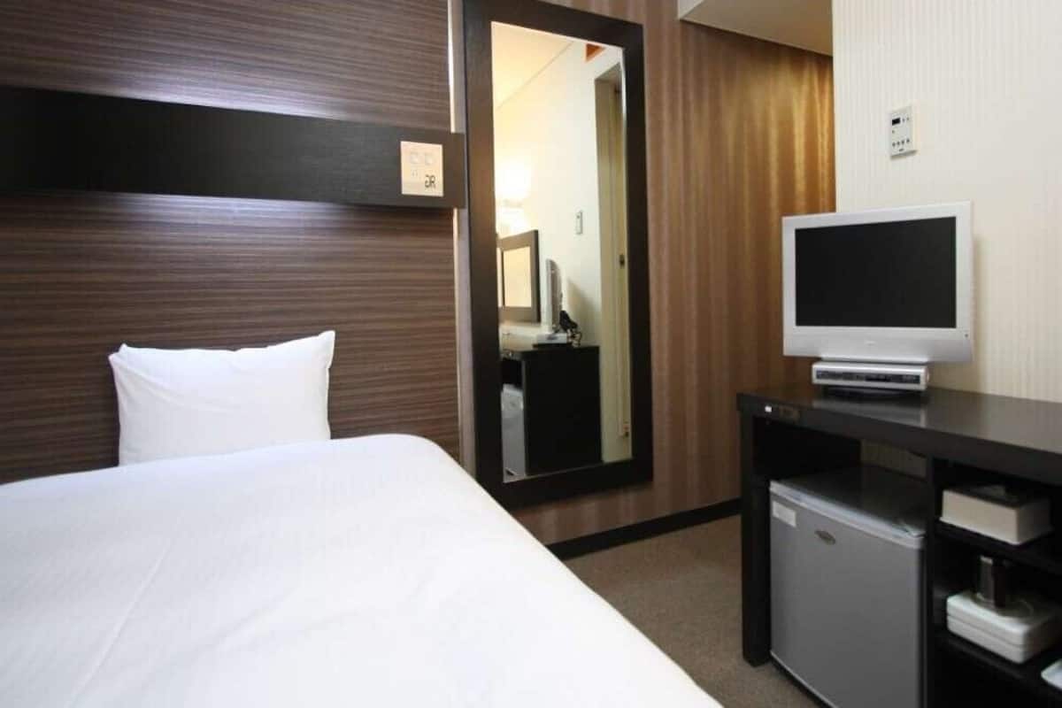 A hotel room with a bed, a small desk with a TV, a mirror, and a wardrobe.