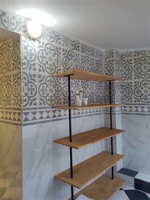A bathroom with patterned tiles, a wooden shelf unit, and a white vase on the countertop.